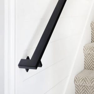 May include: A matte black handrail with a square profile mounted to a white horizontal shiplap wall. The handrail runs diagonally alongside a staircase covered in a light gray and white carpet featuring a repeating herringbone pattern.