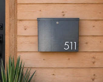 May include: A rectangular charcoal grey metal mailbox mounted on a light brown horizontal wood plank wall. The mailbox features a silver key lock at the top centre and the white house number 511 printed in a clean sans-serif font on the lower right.
