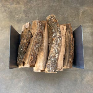Op de afbeelding: Een metalen houtrek met een stapel brandhout erin. Het rek is gemaakt van donker metaal en heeft een rechthoekige vorm. Het brandhout is een mix van bruine en grijze kleuren.