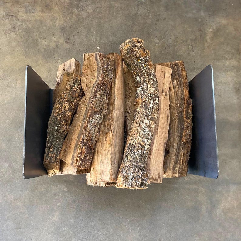 Op de afbeelding: Een metalen houtrek met een stapel brandhout erin. Het rek is gemaakt van donker metaal en heeft een rechthoekige vorm. Het brandhout is een mix van bruine en grijze kleuren.