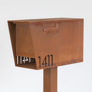 Modern mailbox made of weathered hot rolled steel with a rich rust patina. The laser-cut address number '1411' is displayed on the angled front panel. The mailbox features a minimalist flag and is mounted on a matching steel post. Photographed against a clean white background.