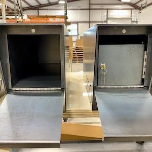 Two raw steel mailboxes displayed side by side in a workshop setting, both with their front doors open. The mailbox on the left shows an open, empty interior, while the one on the right features a secure locking compartment with a hinged door and a key inserted in the lock. Laser-cut address numbers are partially visible on the sides, and shelving, tools, and materials are seen in the background.