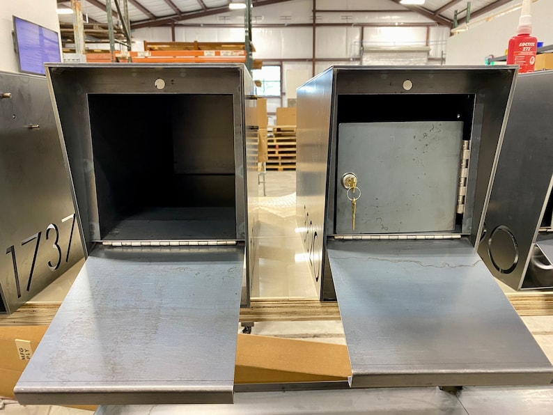 Two raw steel mailboxes displayed side by side in a workshop setting, both with their front doors open. The mailbox on the left shows an open, empty interior, while the one on the right features a secure locking compartment with a hinged door and a key inserted in the lock. Laser-cut address numbers are partially visible on the sides, and shelving, tools, and materials are seen in the background.