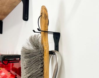 Puede incluir: Un soporte de escoba y mopa de metal negro montado en una pared blanca. El soporte tiene dos ganchos y una barra de metal para sujetar los mangos de la escoba y la mopa. Una escoba con mango de madera y una pala de polvo de metal están colgadas del soporte.