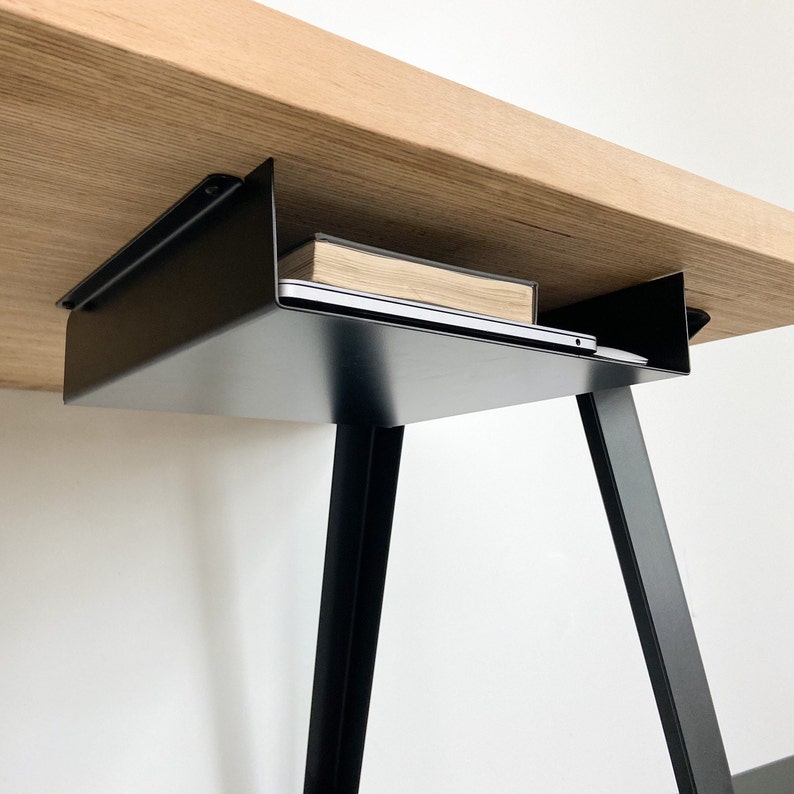 Pu&ograve; includere: Mensola in metallo nero fissata alla parte inferiore di una scrivania in legno. Un laptop e un libro sono appoggiati sulla mensola.