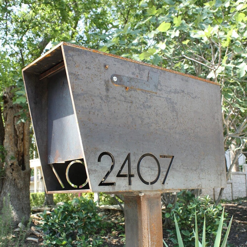 Weathered steel mailbox with a rustic patina finish, featuring laser-cut address numbers '2407' on the angled front panel. The mailbox has a minimalist design with a simple metal flag and is mounted on a matching rusted steel post. Set in a landscaped yard with trees and shrubs in the background.