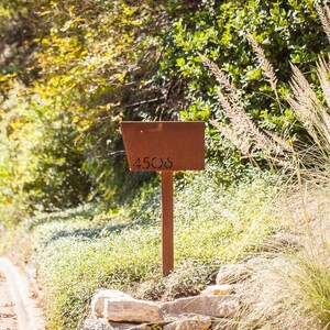 Rust-colored hot rolled steel mailbox with laser-cut address number '4506', mounted on a matching post. The mailbox is situated at the edge of a landscaped driveway, surrounded by lush greenery, ornamental grasses, and a stone border in a sunny, natural setting.
