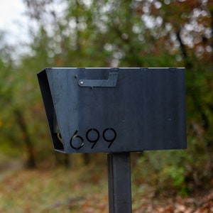 Industrial-style raw steel mailbox with a minimalist metal flag and laser-cut address number '699' on the angled front panel. The mailbox is mounted on a matching steel post and set against a natural, wooded background with soft-focus trees and grass.