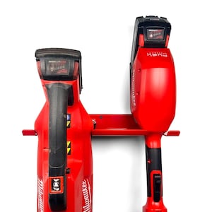 May include: Two red Milwaukee power tools with black handles are mounted on a red metal wall rack. The tools are labeled "FUEL" and have black battery packs.