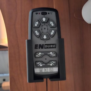 Könnte beinhalten: Eine schwarz-graue Fernbedienung mit der Aufschrift "ENDURO", Richtungstasten und Funktionssteuerungen. Die Fernbedienung ist an einer Holzoberfläche befestigt und hat eine schwarze Handschlaufe.