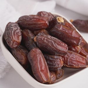 May include: A white ceramic bowl filled with several brown dates. The dates have a wrinkled texture and a glossy sheen. The bowl is set against a white background, with a white cloth visible in the background.
