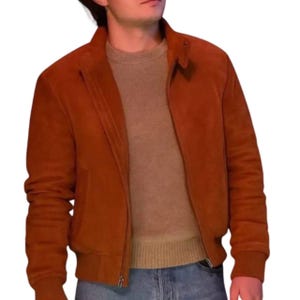 Può includere: Giacca in camoscio color ruggine con zip, colletto e polsini e orlo a coste. La giacca è indossata sopra un maglione beige e jeans blu. La giacca ha un classico stile bomber.