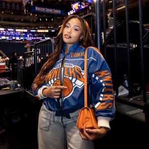 Op de afbeelding: Een blauw en oranje New York Knicks jack met het logo en de letters van het team. De jas heeft witte manchetten en biezen. Een oranje handtas met een lange riem wordt in de hand gehouden. Achtergrond van een basketbalarena.