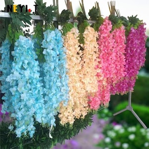 May include: Artificial wisteria vines in shades of light blue, peach, pink, and purple are displayed. The flowers hang from a metal structure with green leaves. The image is brightly lit, highlighting the vibrant colours of the floral arrangements.
