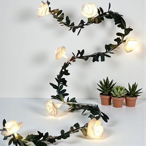 Puede incluir: Una guirnalda de luces decorativa con bombillas blancas en forma de rosa y hojas de color verde oscuro. Las luces están dispuestas en un patrón sinuoso, con tres pequeñas suculentas en macetas en el fondo. Para interiores.