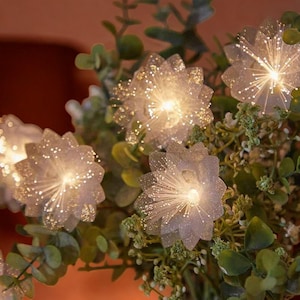Op de afbeelding: Close-up van een bloemenlichtslinger. De slinger heeft heldere, bloemvormige lampjes met een sprankelende, glitterende textuur. De lampjes zijn afgewisseld met groen blad, waardoor een zacht, gloeiend effect ontstaat. De lampjes zijn verlicht en creëren een warme sfeer.