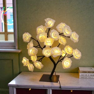 Op de afbeelding: Een decoratieve tafellamp in de vorm van een boom, met witte roosvormige lampjes op zwarte takken. De lamp heeft een vierkante voet en een netsnoer. De rozen geven een warme, uitnodigende gloed.