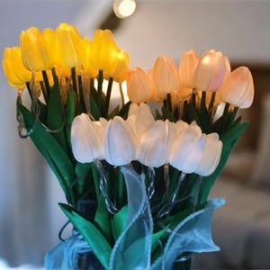 Puede incluir: Un ramo de tulipanes artificiales iluminados en amarillo, melocotón y blanco. Las flores artificiales tienen tallos y hojas verdes, con una cinta azul en la base. Los tulipanes están iluminados desde dentro, creando un brillo suave.
