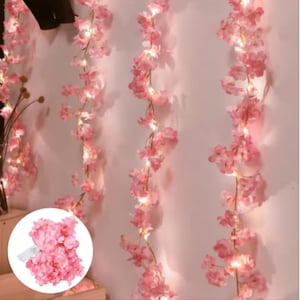 Può includere: Stringa luminosa di fiori di ciliegio rosa. Le luci sono disposte lungo un filo flessibile, con ogni fiore illuminato da un piccolo LED. Le luci sono disposte verticalmente contro un muro rosa chiaro, creando un'atmosfera morbida e romantica.