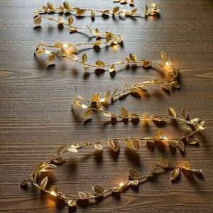 May include: Gold-colored leaf garlands with small, warm white lights. The garlands are arranged in a flowing pattern on a wooden surface. The leaves are detailed and create a decorative, natural look. Ideal for home decor or event decorations.