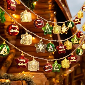 Puede incluir: Una cadena de luces navideñas con varios adornos. Las luces incluyen cajas de regalo, estrellas y adornos redondos en rojo, verde y dorado. Algunos adornos tienen un diseño de árbol de Navidad o la inscripción "Merry Christmas".