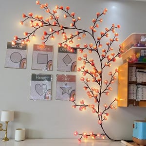 Puede incluir: Una rama decorativa iluminada de flores de cerezo con flores rosas y luces blancas cálidas. La rama está montada en una pared blanca, junto a tarjetas educativas de formas geométricas con etiquetas.