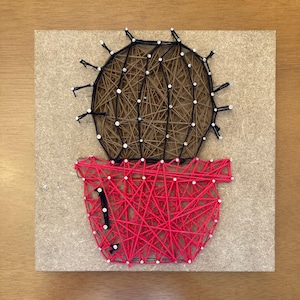 以下が含まれることがあります： 赤い鉢に入ったストリングアートのサボテンが正方形の木の板に飾られています。サボテンは茶色で、黒いアウトラインと白いアクセントがあります。鉢は赤色で、この作品は糸と釘で作られています。