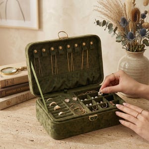 Könnte beinhalten: Geöffnete olivgrüne Samt-Schmuckschatulle mit goldfarbener Hardware. Die Schatulle ist mit verschiedenen goldenen Halsketten, Ringen und Ohrringen gefüllt. Eine Hand legt einen Perlenohrring hinein. Die Schatulle ist auf einer hellen Oberfläche geöffnet.
