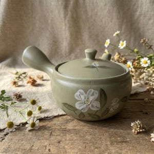 May include: A sage green ceramic soup bowl with a lid and handle. The bowl is decorated with white floral designs and green leaves. The bowl is sitting on a wooden surface.