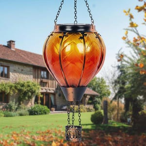 May include: Hanging solar lantern with a warm orange and yellow glass globe, resembling a hot air balloon. The lantern has a black metal frame and chain, with a decorative basket at the bottom. Ideal for garden decor.