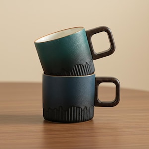 May include: Two ceramic mugs stacked. The top mug is teal, the bottom is navy blue. Both have dark brown square handles and textured, ridged detailing near the base. The top mug's interior is off-white. Ideal for tea or coffee.