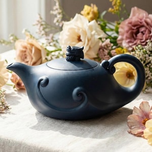 May include: A navy blue ceramic teapot with a dragon-shaped lid handle and a curved spout. The teapot features a decorative swirl design on the body and a large, rounded handle. The background includes flowers.