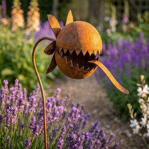 May include: A quirky, rusted metal garden ornament resembling a flower with a spherical head and a toothed mouth. The sculpture has a long, curved stem and a tongue-like projection. The background shows a garden with purple flowers.