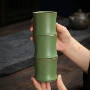 May include: Three-piece green ceramic tea cup set. The cups are stacked vertically, resembling bamboo stalks. Each cup has a slightly flared rim and a subtle gold-colored band. The set is held by a person's hands, with a blurred background.