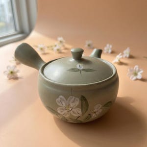 Peut inclure: Une théière en céramique vert sauge avec une anse et un couvercle. La théière est ornée de motifs floraux blancs et de feuilles vertes. Le couvercle comporte un petit bouton et un motif floral peint. La théière est conçue pour infuser le thé.