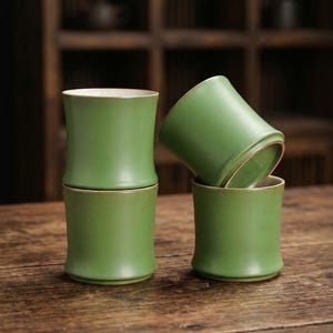 May include: Four green ceramic cups stacked on a wooden surface. The cups have a smooth, matte finish with a slightly flared base and a contrasting light-colored rim. The cups are arranged in a visually appealing composition.