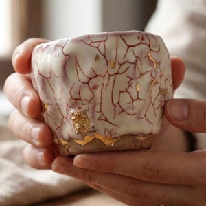 May include: A handcrafted ceramic cup with a unique crackle glaze pattern in red and white. The cup features gold leaf accents and a textured surface, showcasing a blend of traditional craftsmanship and modern design.