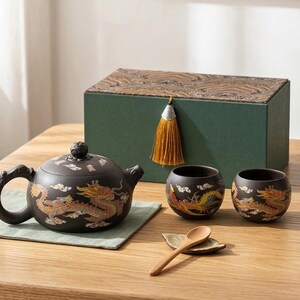 May include: A black ceramic tea set featuring dragon motifs in gold, red, and yellow. The set comprises a teapot, two cups, a wooden spoon, and a small leaf-shaped dish. A green box with a decorative lid and tassel is in the background.