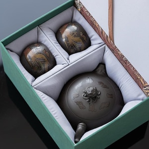 May include: A dark grey tea set presented in a green box. The set includes a teapot and two cups, each decorated with a gold phoenix design. The teapot has a decorative lid and spout. The box is lined with white fabric.