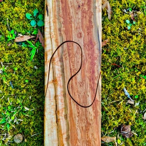 Könnte beinhalten: Ein rechteckiges Holzbrett mit einer geschnitzten, geschwungenen Linie. Das Holz hat ein natürliches, rötlich-braunes Maserungsmuster. Das Brett liegt vor einem Hintergrund aus grünem Moos und kleinen Pflanzen.