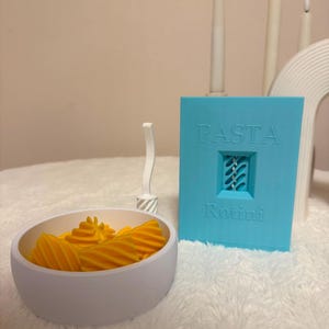 Könnte beinhalten: Ein hellblauer rechteckiger Behälter mit der Aufschrift PASTA Rotini mit einem mittigen Ausschnitt, einem kleinen weißen Werkzeug und einer weißen Schale mit gelben spiralförmigen Nudeln. Der Behälter hat geprägten Text und eine sichtbare Struktur.