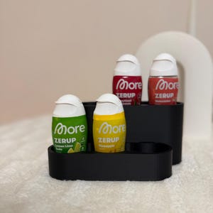 Könnte beinhalten: Ein schwarzer, gestufter Kunststoff-Organizer mit vier kleinen Quetschflaschen der Geschmacksverstärker More Zerup. Die Flaschen sind mit Lemon Lime Soda, Pineapple und zwei weiteren Sorten beschriftet und haben weiße Klappverschlüsse.