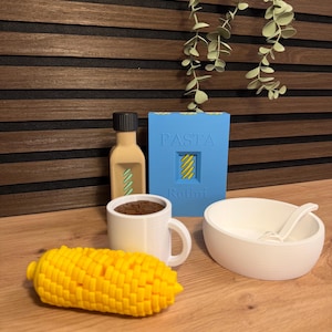 Könnte beinhalten: Eine Sammlung von Miniatur-Lebensmitteln, bestehend aus einem gelben Maiskolben, einer weißen Tasse mit braunem Inhalt, einer beigen Flasche mit schwarzem Deckel, einer blauen Schachtel mit der Aufschrift PASTA Rotini und einer weißen Schale mit Löffel.