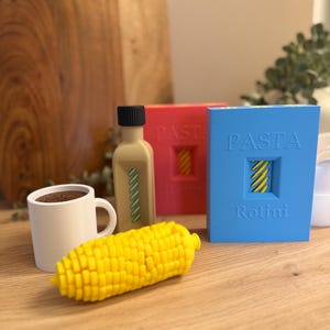 Könnte beinhalten: Eine Sammlung von 3D-gedruckten Objekten, bestehend aus einem weißen Becher mit braunem Inhalt, einer beigen Flasche mit schwarzem Deckel, einem gelben Maiskolben und zwei rechteckigen Schachteln mit der Aufschrift PASTA Rotini in Blau und Rot.
