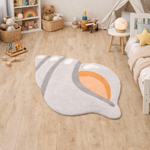 May include: A seashell-shaped rug in a child's room. The rug is primarily light gray with gray stripes and an orange interior. The room has a white bed, a small table, and decorative wall hangings, including clouds and a sun.