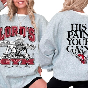 Könnte beinhalten: Ein hellgraues Sweatshirt mit Rundhalsausschnitt und einem Grafikdruck auf der Vorderseite mit dem Text LORD'S GYM, THE SIN OF THE WORLD und BENCH PRESS THIS! sowie einer Abbildung einer Figur mit Kreuz. Hinten steht HIS PAIN YOUR GAIN.