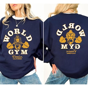 Pode incluir: Camisola azul marinho com gola redonda branca. A frente apresenta um gráfico dourado e branco de um gorila levantando pesos, as palavras "WORLD GYM" e "Venice, Califórnia". A parte de trás tem um design semelhante.