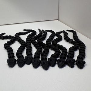 May include: A collection of black articulated snake toys, each with a segmented body and a detailed head. The snakes are arranged in a group, showcasing their flexible design and dark color against a white background.