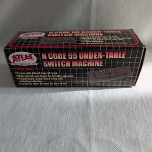 May include: A black and red box for an Atlas N Code 55 Under-Table Switch Machine. The box features the product name in large, bold letters, along with the Atlas Model Railroad Co. logo. The box also includes product details and specifications.