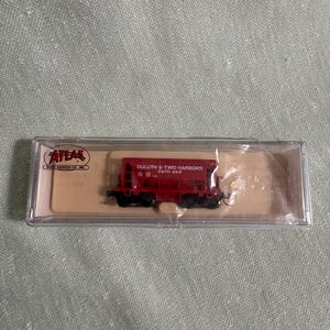 May include: A red model train car with black wheels and the text "DULUTH & TWO HARBORS" and "D&TH 262" printed on the side. The model is in a clear plastic case with the "ATLAS" logo.